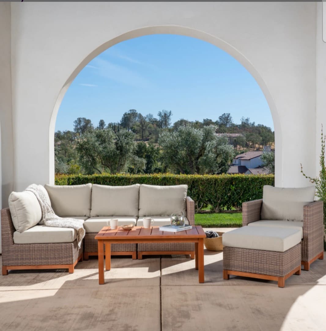 Wicker patio furniture with beige cushions and wooden coffee table under a white archway.
